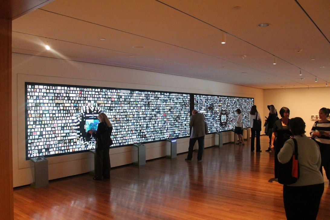 An interior view of a digital art gallery with visitors viewing a large wall-sized digital display composed of many small images or videos.